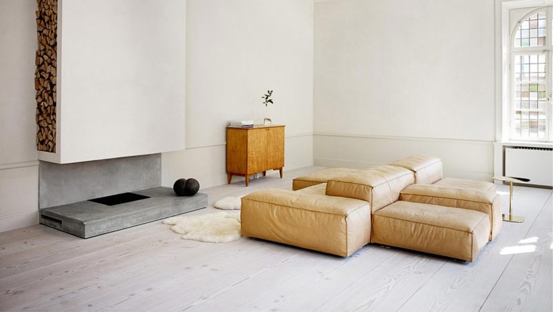 Phòng khách tối giản với tông màu trung tính, sàn gỗ sáng và sofa da mô-đun linh hoạt. Lò sưởi bê tông kết hợp kệ gỗ tạo điểm nhấn thô mộc, mang lại vẻ ấm cúng và tự nhiên.
